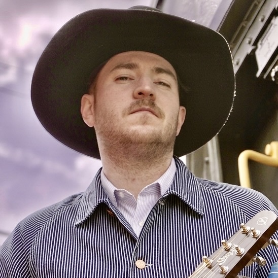 Close-up portrait of Austin T. O'Keefe, Canadian traditional country musician, wearing a black cowboy hat and striped denim jacket, holding an acoustic guitar against a moody sky background.