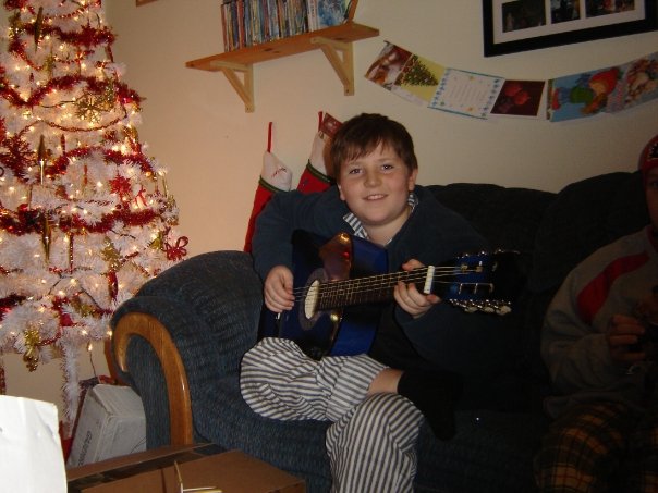 A young boy smiling and holding a blue acoustic guitar in a playing position while sitting in front of a white Christmas tree. Austin T. O'Keefe starting his self-taught musical journey with a blue guitar.