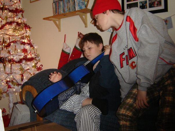 A young boy in striped pajamas sitting on a blue armchair, looking down intently at a new blue acoustic guitar on Christmas morning.