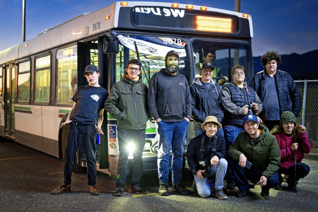 Austin T. O'Keefe kneeling with a group of transit enthusiasts and volunteers in front of a vintage BC Transit bus at dusk.