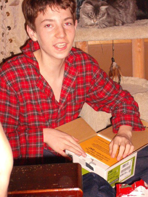 A teenage boy in red plaid pajamas excitedly unboxing a new JVC Everio S camcorder on the floor.