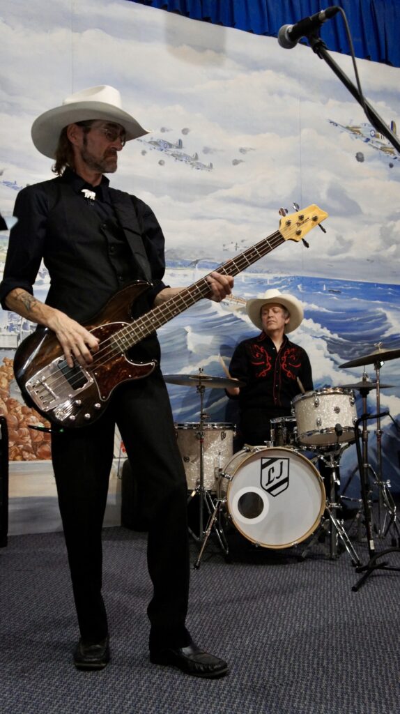 Lorne Gelowitz playing a sunburst precision bass and Lonnie James on drums during a live performance by the Nostalgia Cowboys. They are wearing white cowboy hats and western shirts in front of a mural backdrop. The Sound of the Texas Troubadour: Bridging the Gap from Alberta to the 1970s. Ernest Tubb and Western Swing