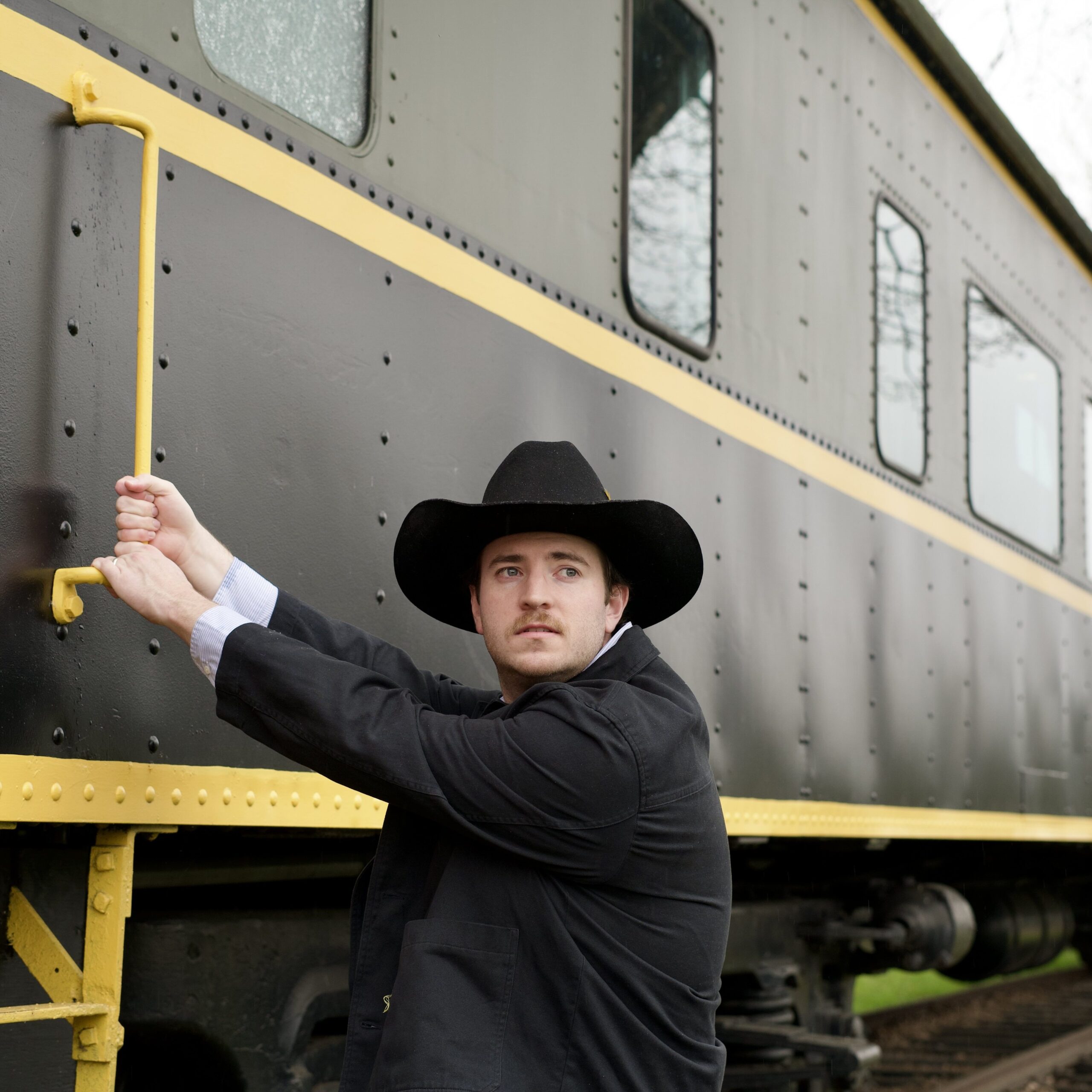 Austin T. O'Keefe in a black cowboy hat and jacket, standing beside a vintage dark green and yellow train car on a railway track.